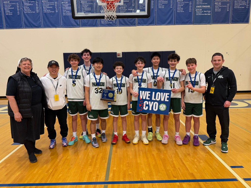 A group of young basketball players wearing green and white uniforms, standing together in a gymnasium with a victory plaque and a sign that says 'WE LOVE CYO'. Coaches and a female team member are also present in the photo.