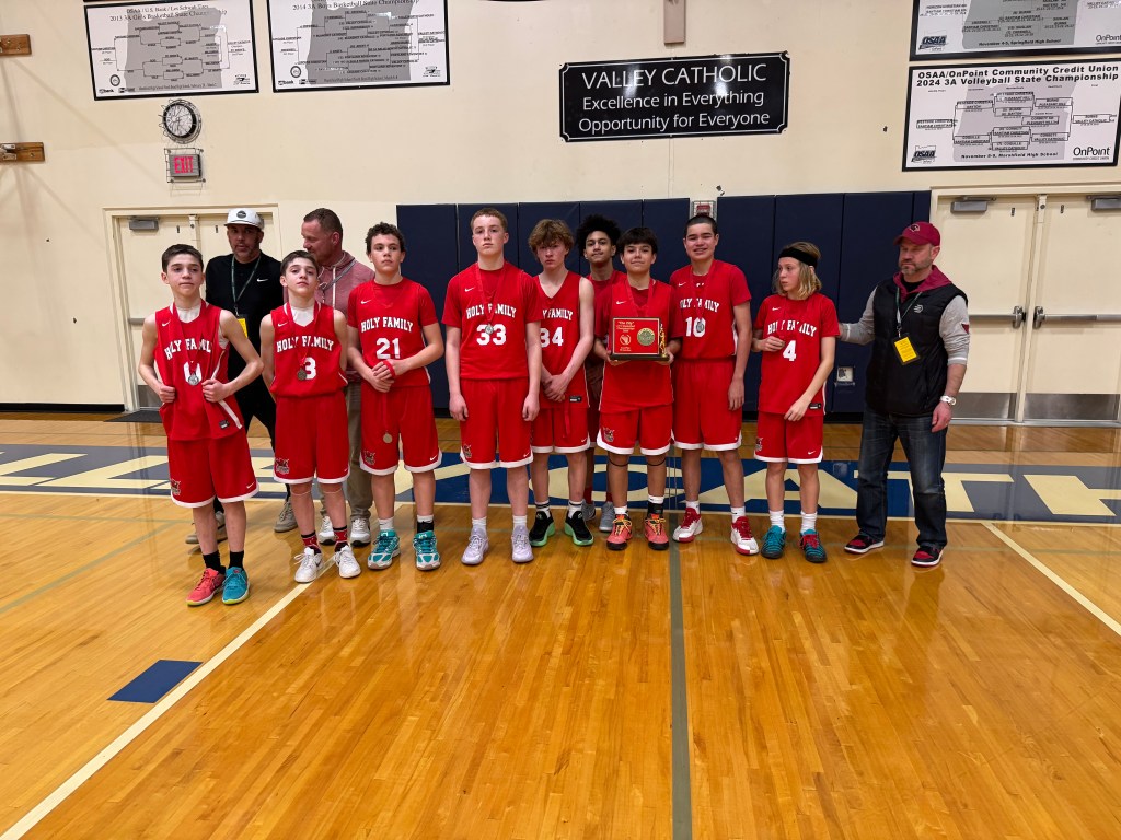 A youth basketball team in red uniforms poses for a group photo in a gymnasium, holding a trophy. They are standing on a wooden floor with sports banners in the background.